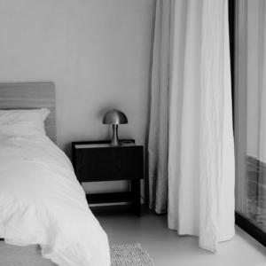 Calm black-and-white bedroom set up for a Sunday routine, with a freshly made bed in white linen, a dark wooden bedside table, a silver mushroom lamp and long linen curtains by a window.