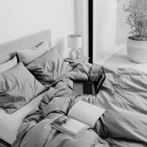 Unmade bed in soft morning light with grey linen, an open magazine and a laptop, beside a sculptural lamp and a potted bamboo plant, for an article on becoming a morning person when you’re not one.