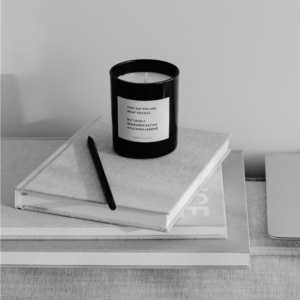 Black-and-white photo of a minimalist desk with a candle resting on a closed linen notebook and a pencil, next to a laptop, for an article about setting daily goals that create progress.