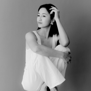 Black-and-white portrait of a woman in a white top and trousers sitting with her knees drawn up, one hand in her hair, gazing thoughtfully to the side, for an article about why you feel lazy and unmotivated.