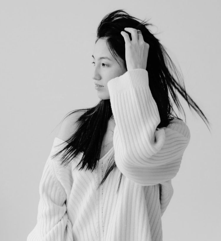 Black and white photo of a woman with long dark hair running her hand through it, wearing an oversized off-the-shoulder ribbed sweater against a plain light wall