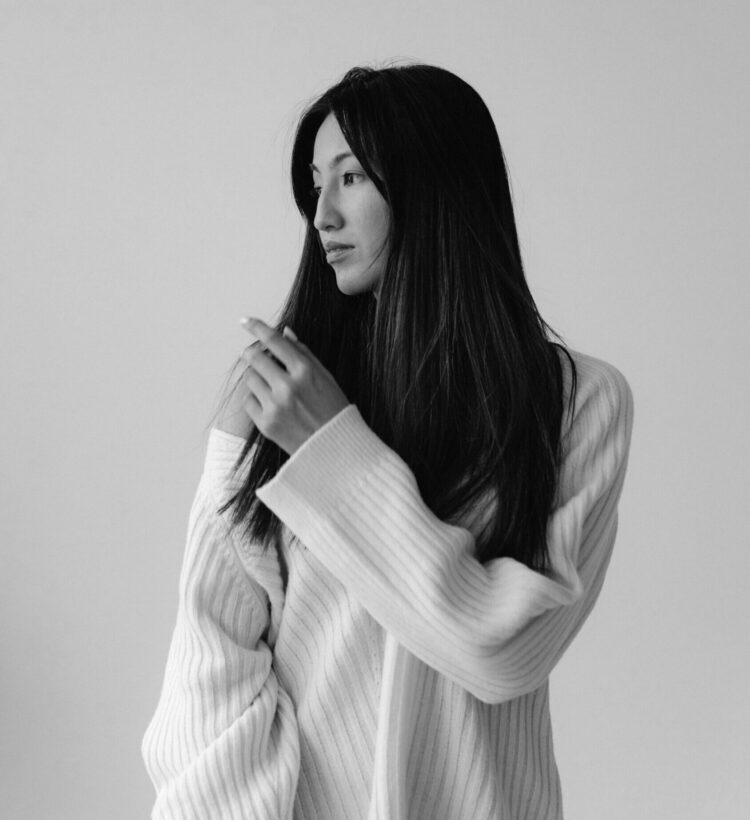 Black and white photo of a woman with long dark hair in an oversized ribbed sweater, looking away in quiet thought