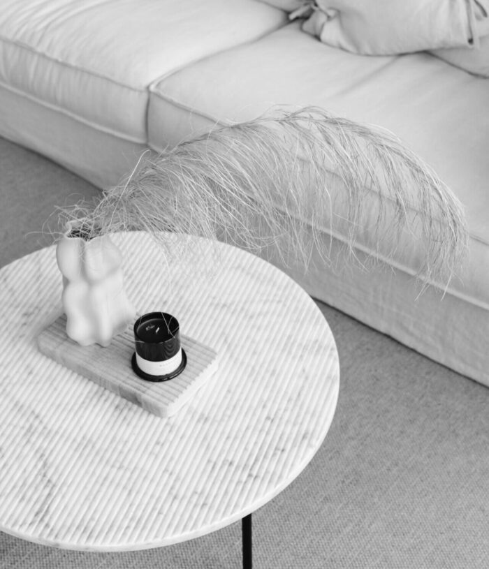 Black and white photo of a round marble coffee table with dried pampas grass, a candle and a sculptural vase in front of a linen sofa, for the Foundations section